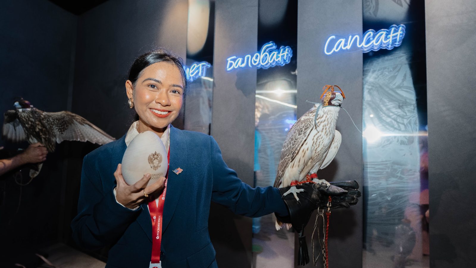 Siti Zulaikha holding a falcon on a gloved hand and a commemorative egg, in front of illuminated display panels.