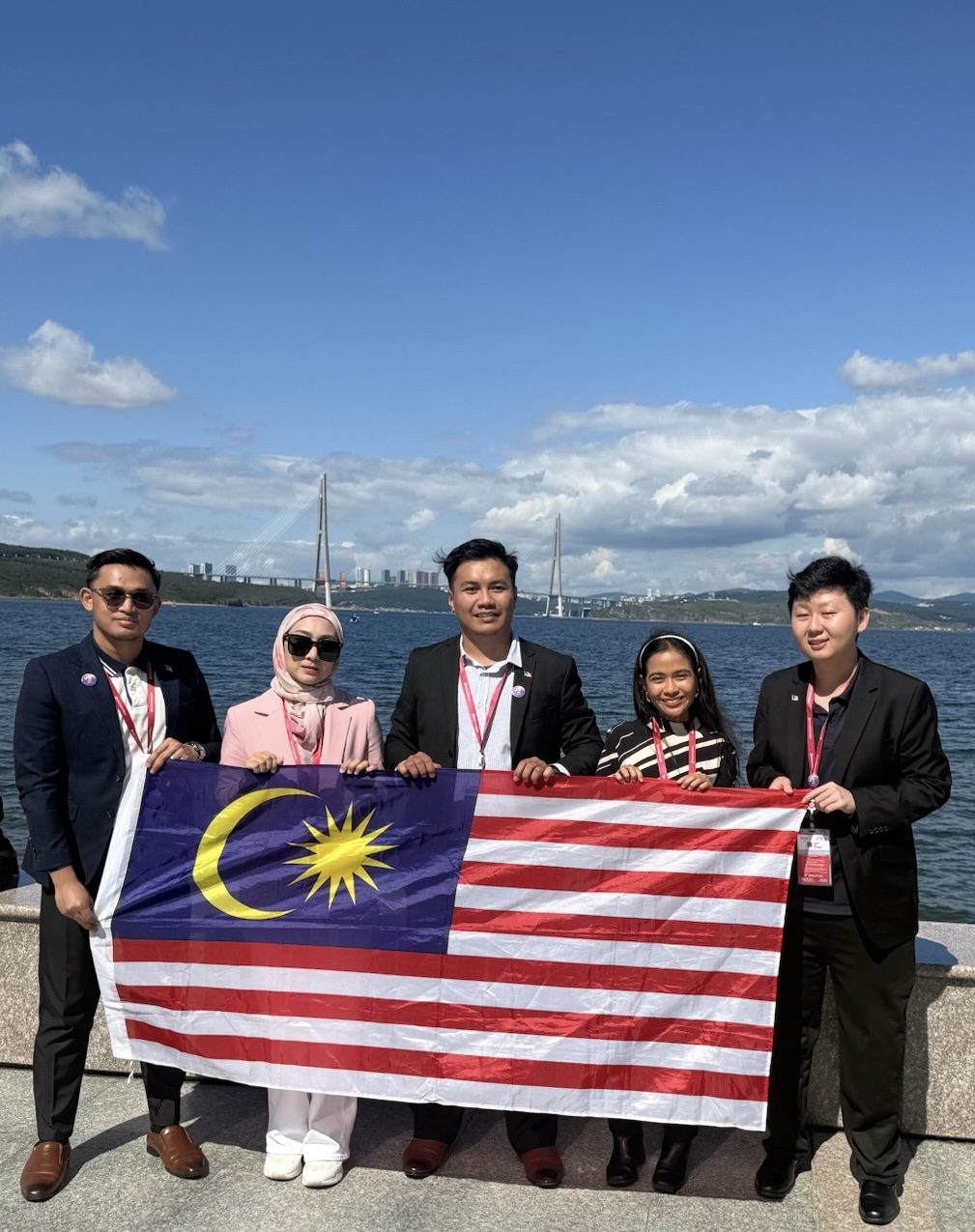 A group of Malaysian delegates holding the Malaysian flag beside the Vladivostok waterfront, with a bridge in the background.